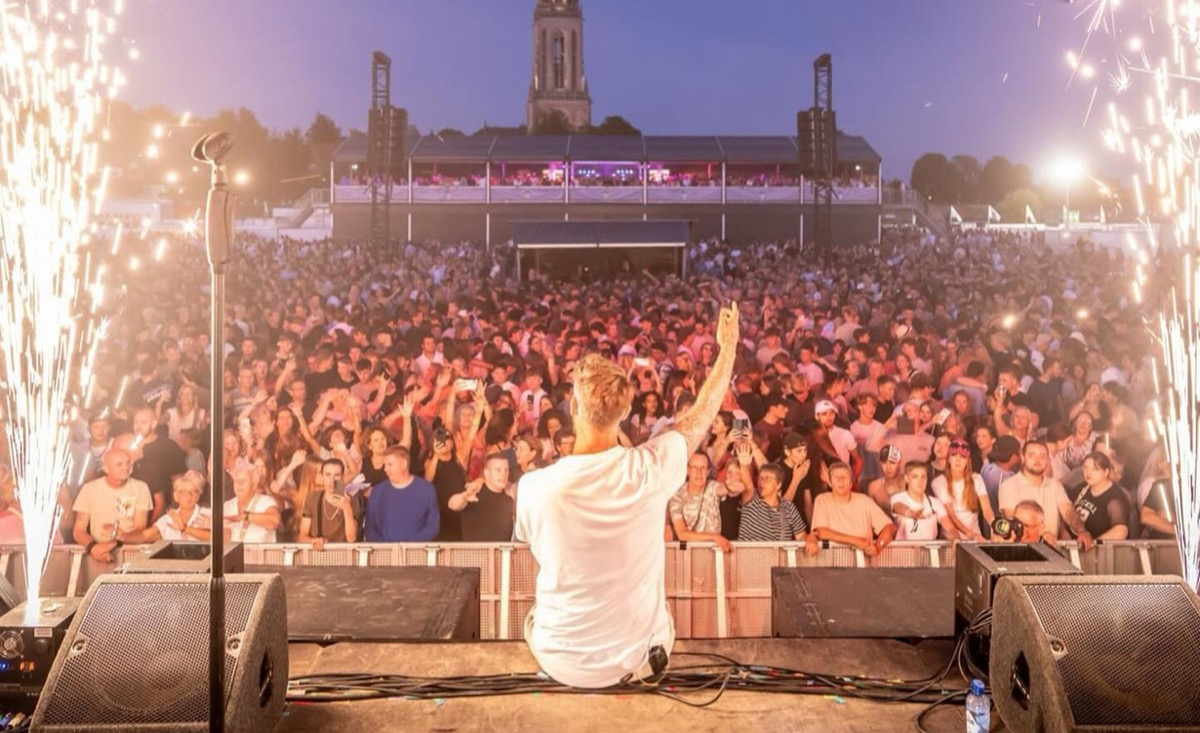 Marcel Swerissen zit op de rand van een festivalpodium met zijn arm omhoog, voor een terrein vol publiek met vuurwerkfonteinen aan weerszijden.