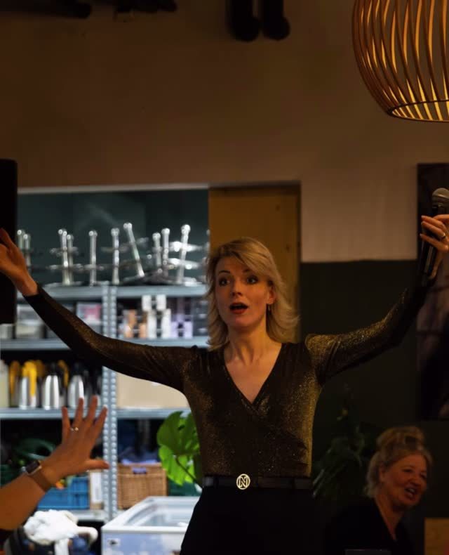 Een zangeres staat tussen de tafels met haar armen gespreid, gasten om haar heen.